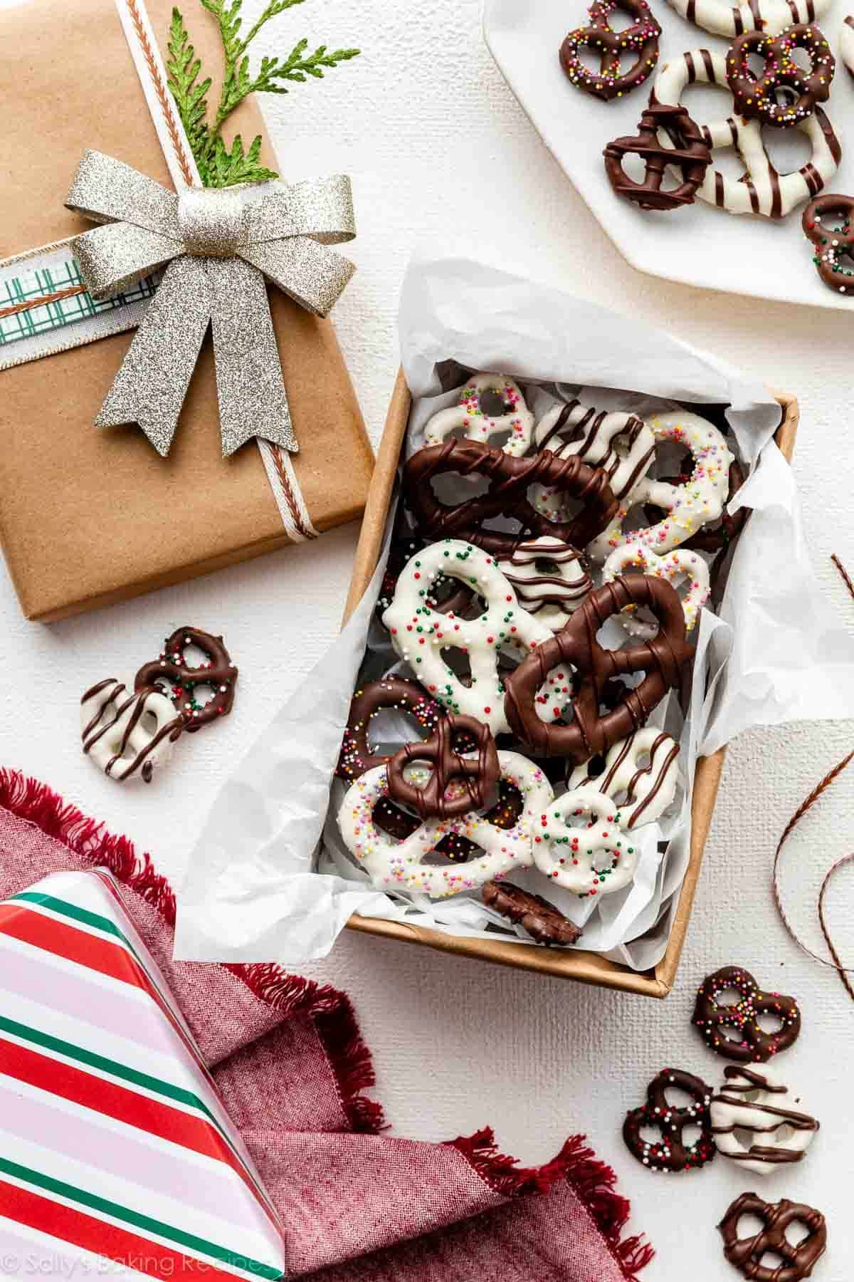 box of various chocolate covered pretzels including some with white chocolate, chocolate drizzles, and sprinkles surrounded by wrapped Christmas gifts.
