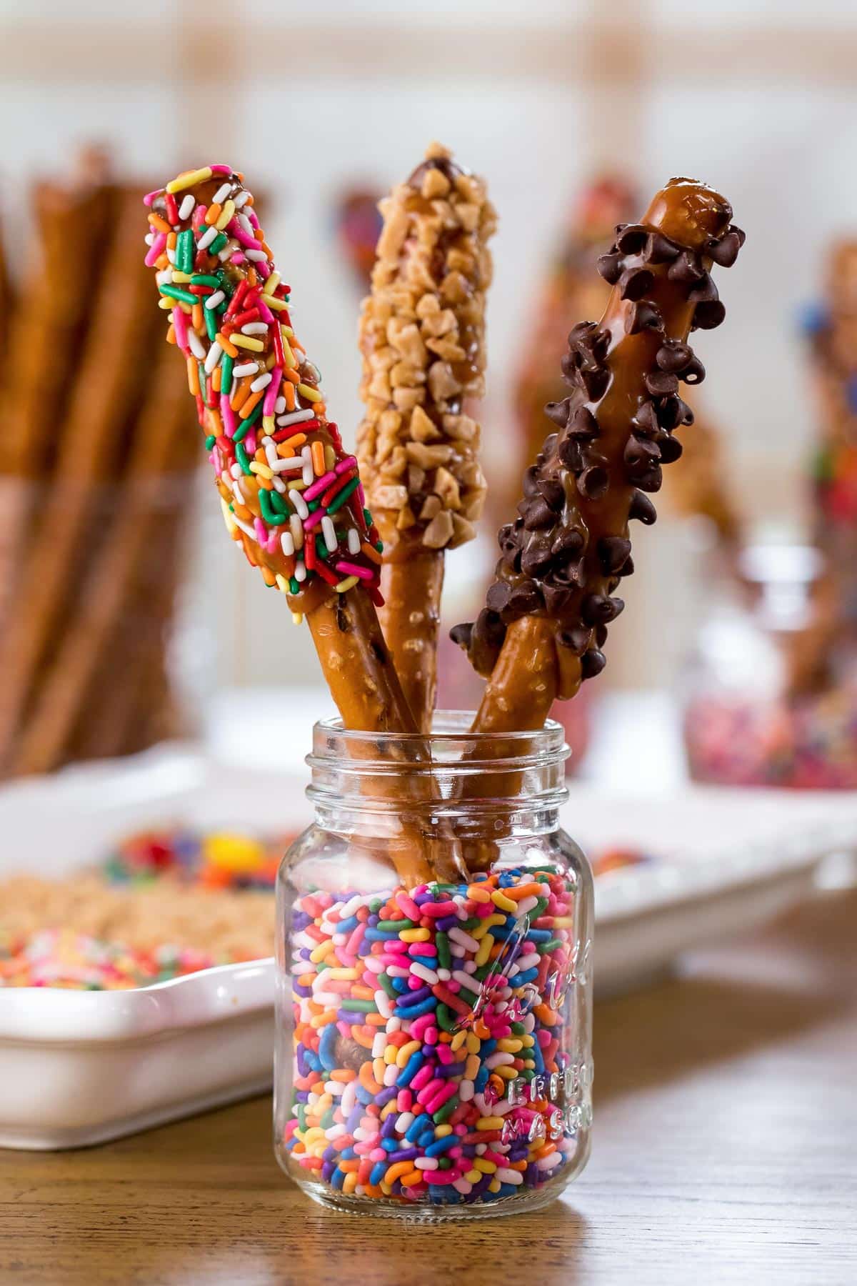 3 pretzel rods dipped in caramel and various toppings standing up in a glass jar with sprinkles