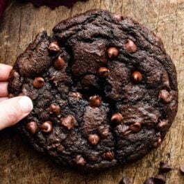 one giant double chocolate cookie on wooden cutting board with hand pulling broken half away.