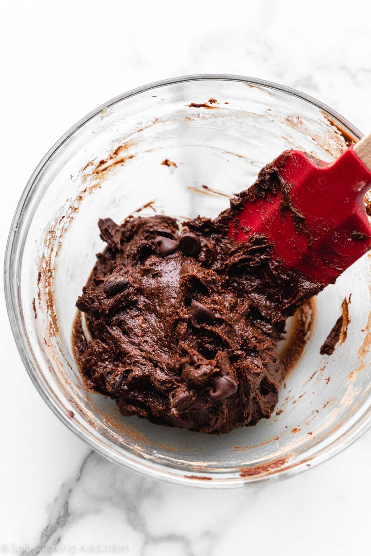 small amount of chocolate cookie dough in glass bowl with red spatula.