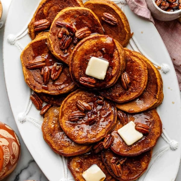 pumpkin pancakes with slices of butter and maple pecan syrup on top sitting on white platter.
