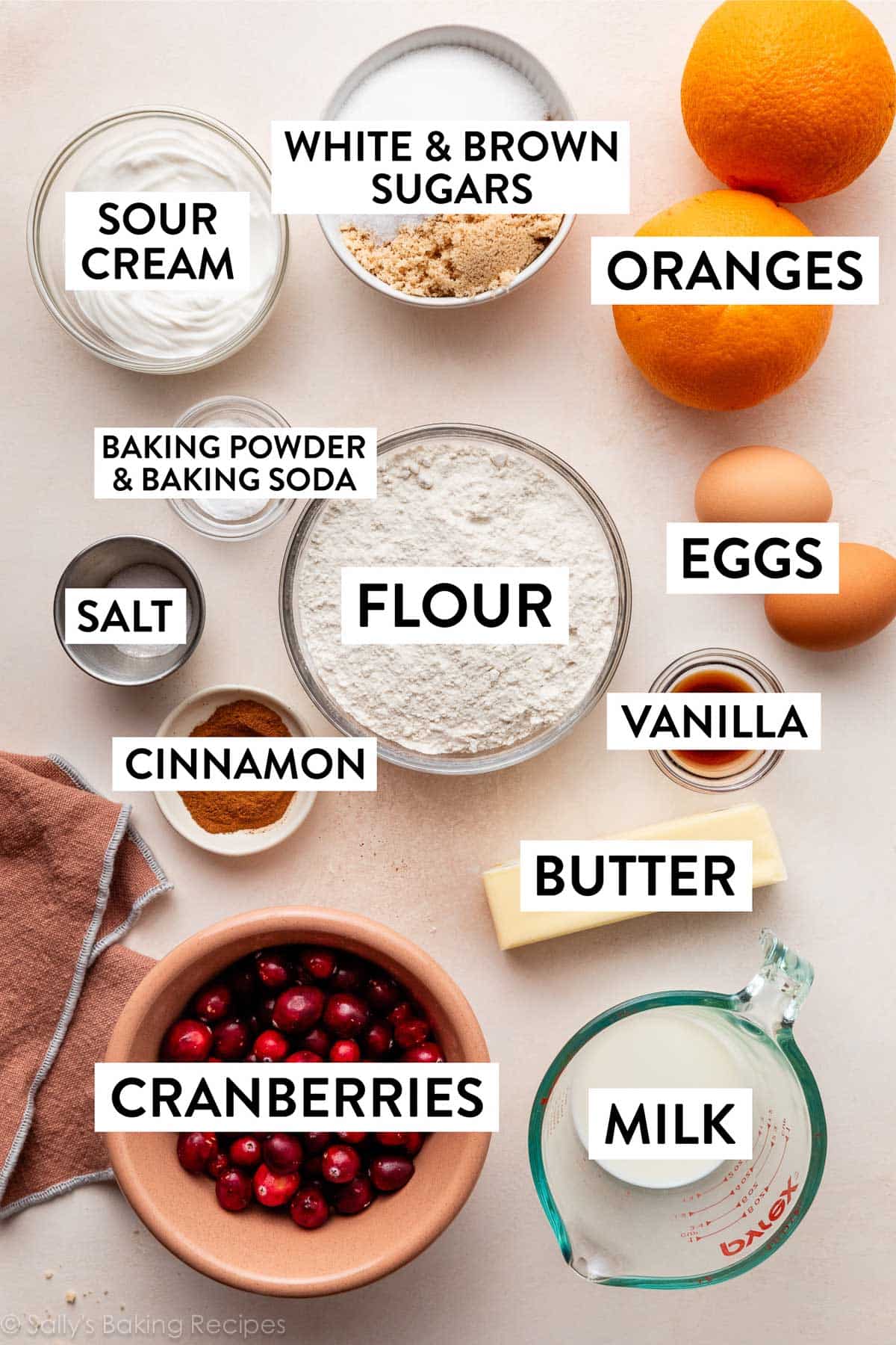 ingredients on peach-cream surface including eggs, vanilla, butter, milk, flour, sugars, and oranges.