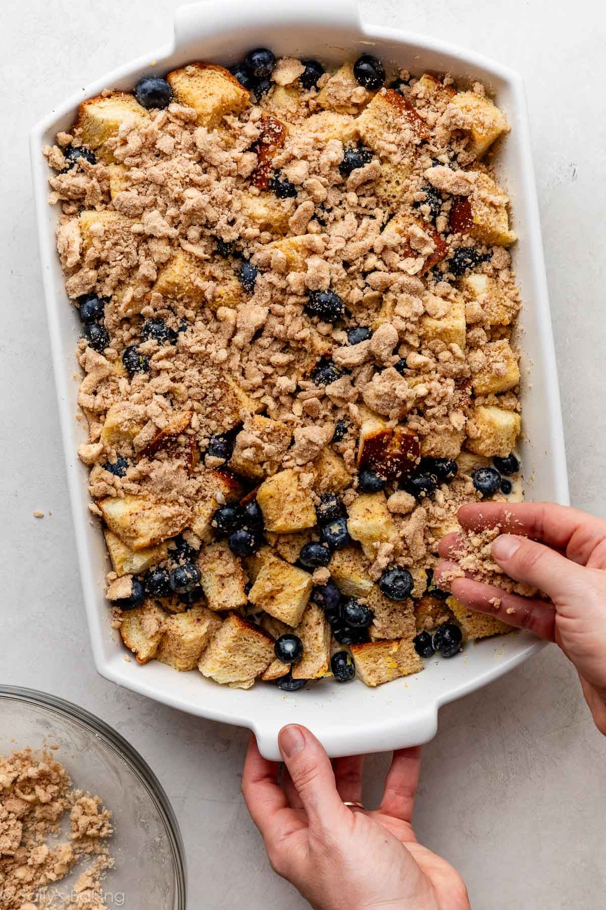 hands adding crumb topping to bread and blueberries in casserole dish.