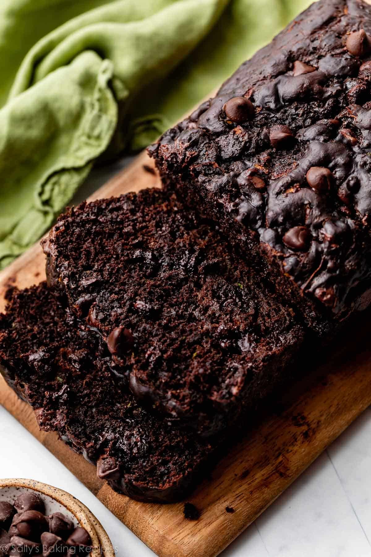 2 slices and the loaf of chocolate zucchini bread on wooden cutting board with green linen in the background.