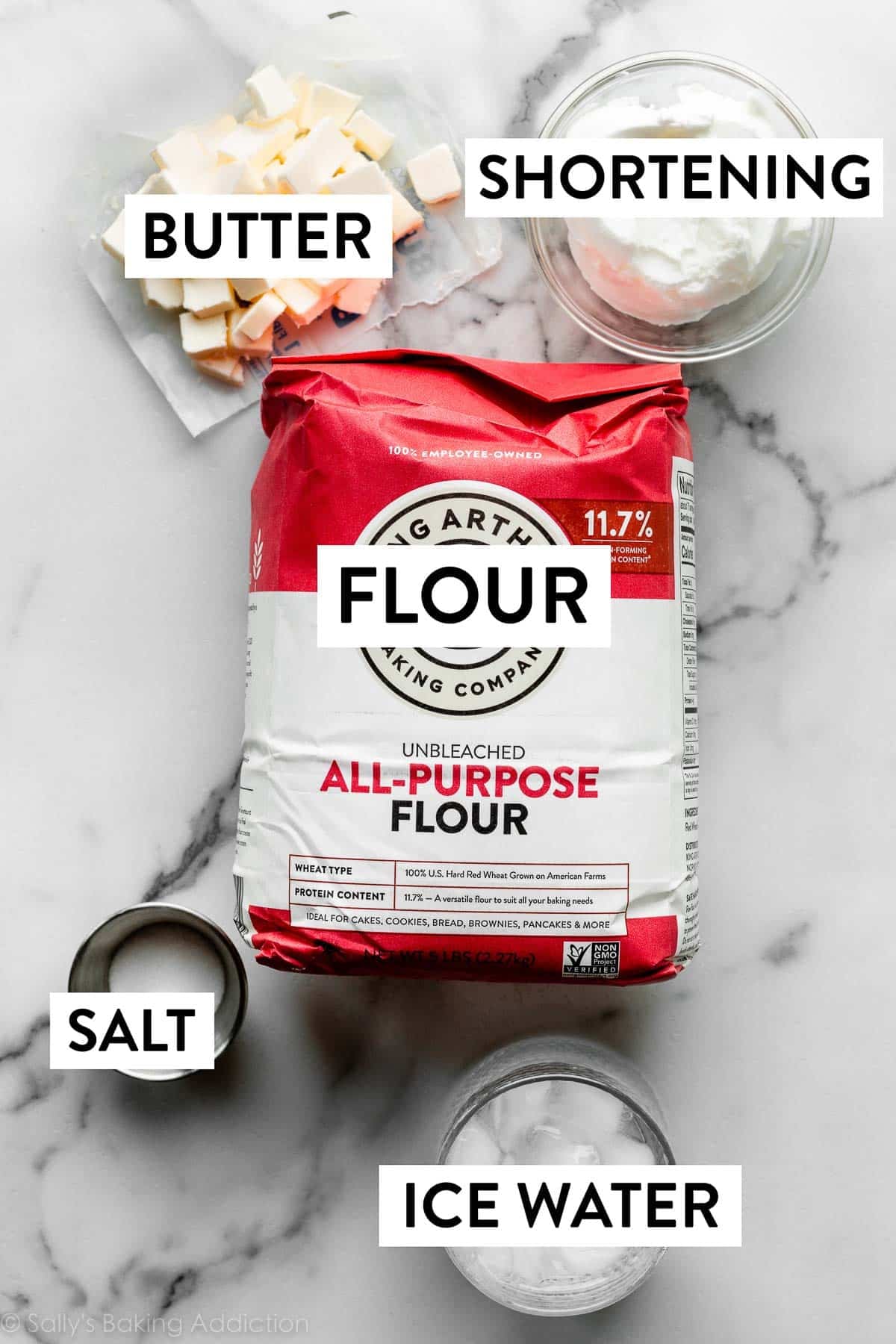 ingredients on marble counter including flour, shortening, butter, salt, and a cup of ice water.