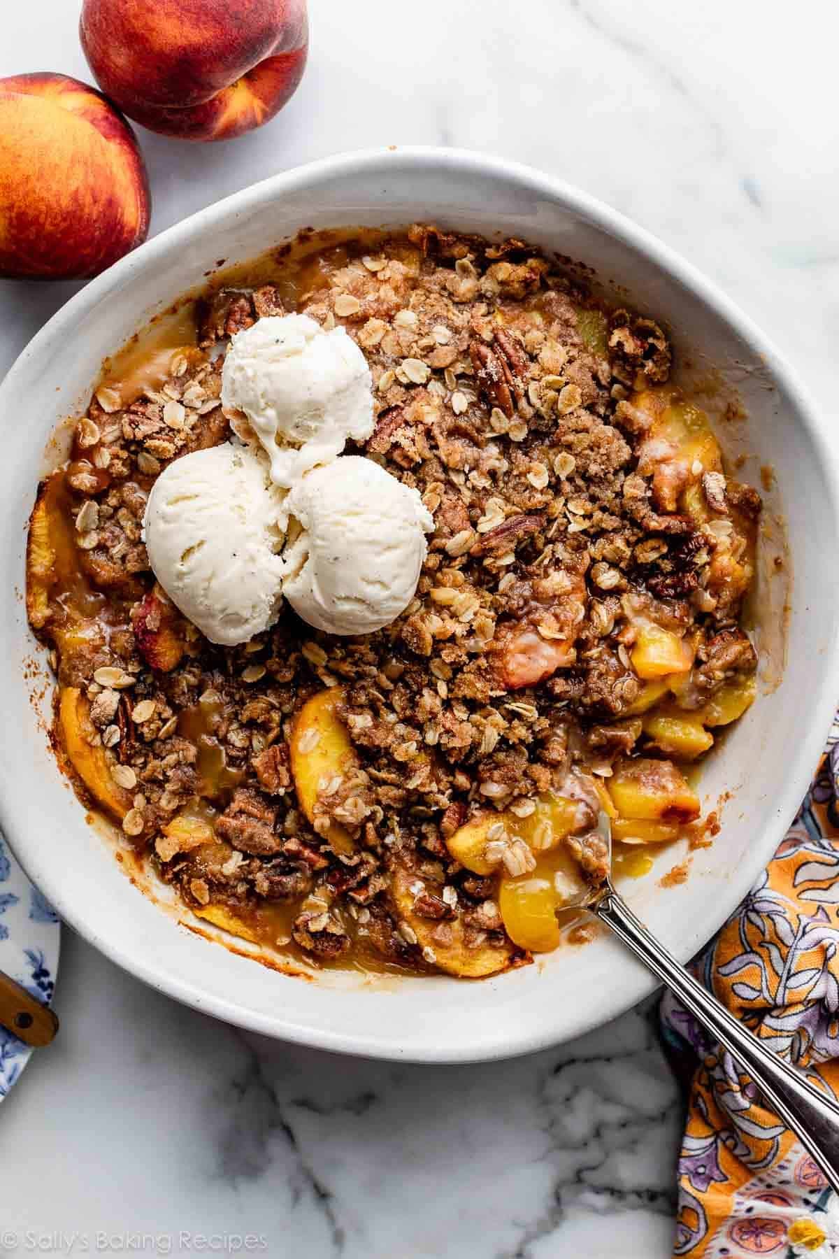 peach pecan crisp in white round baking dish with 3 vanilla ice cream scoops on top.