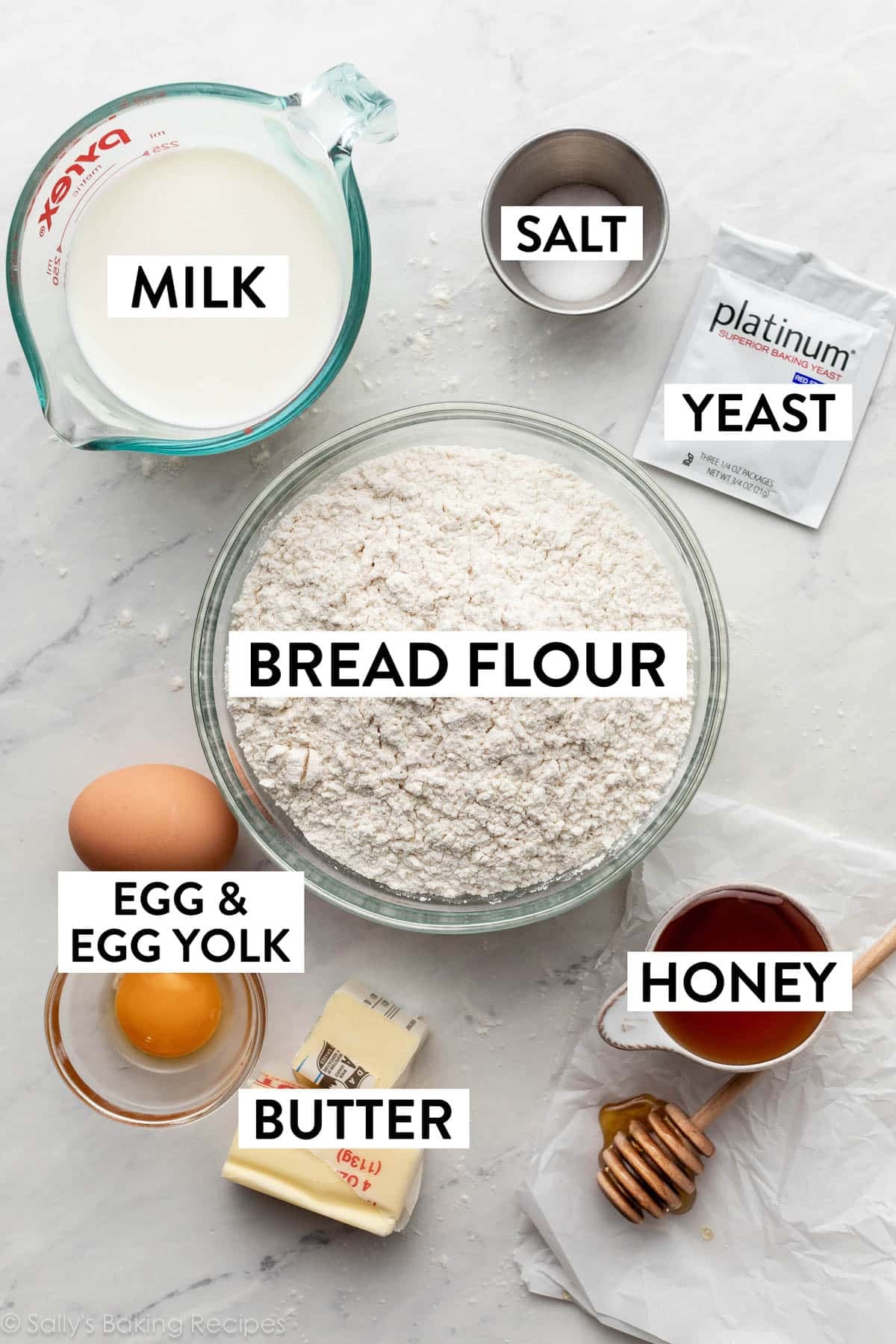 ingredients on counter including a packet of yeast, bowl of bread flour, milk, salt, honey, butter, 1 egg, and egg yolk.