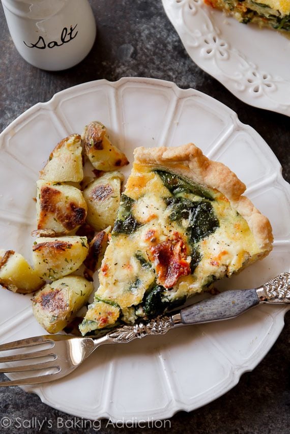 slice of goat cheese spinach sun-dried tomato quiche with breakfast potatoes on a white plate with a fork
