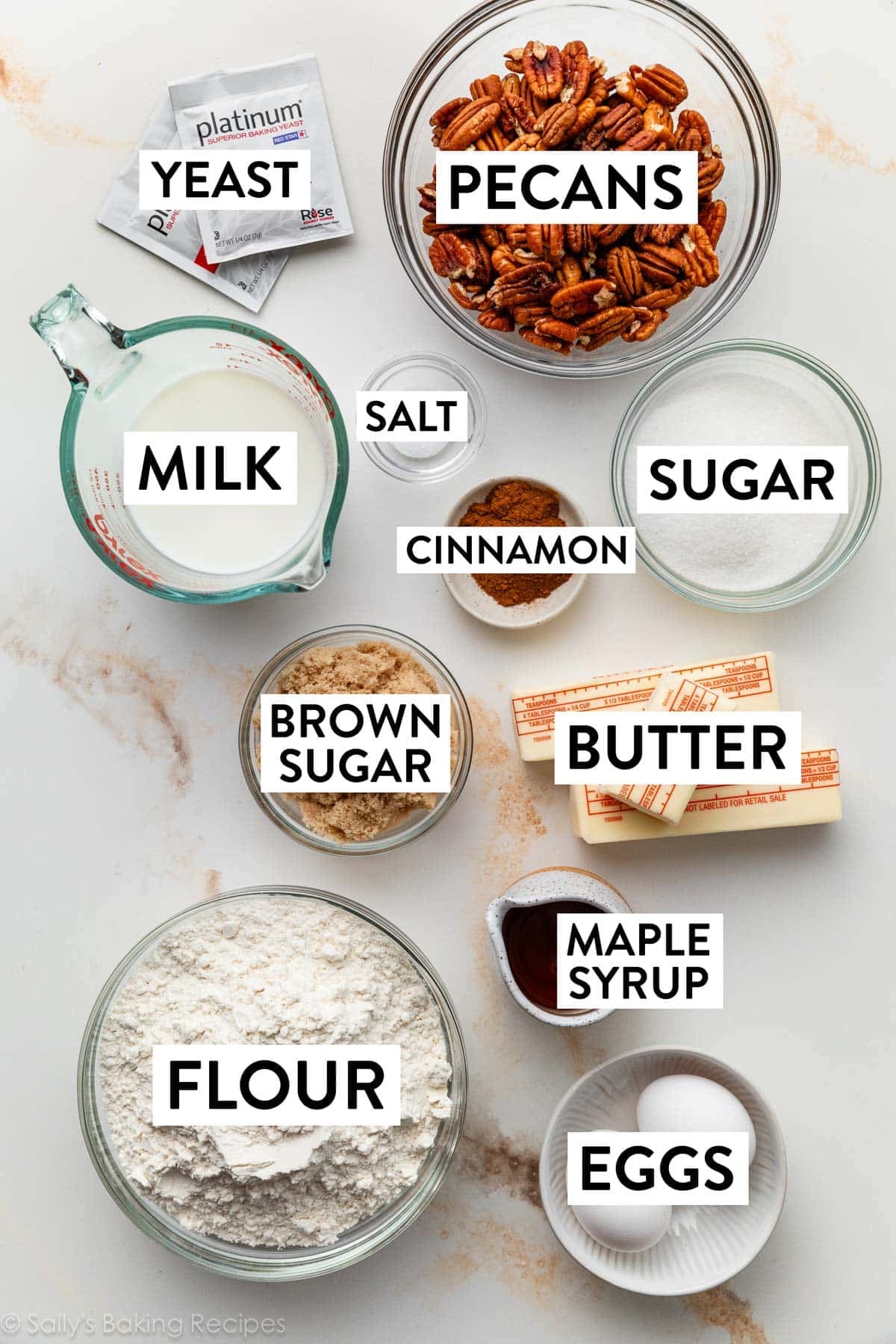 ingredients on surface including flour, eggs, maple syrup, butter, brown sugar, cinnamon, yeast, and milk.