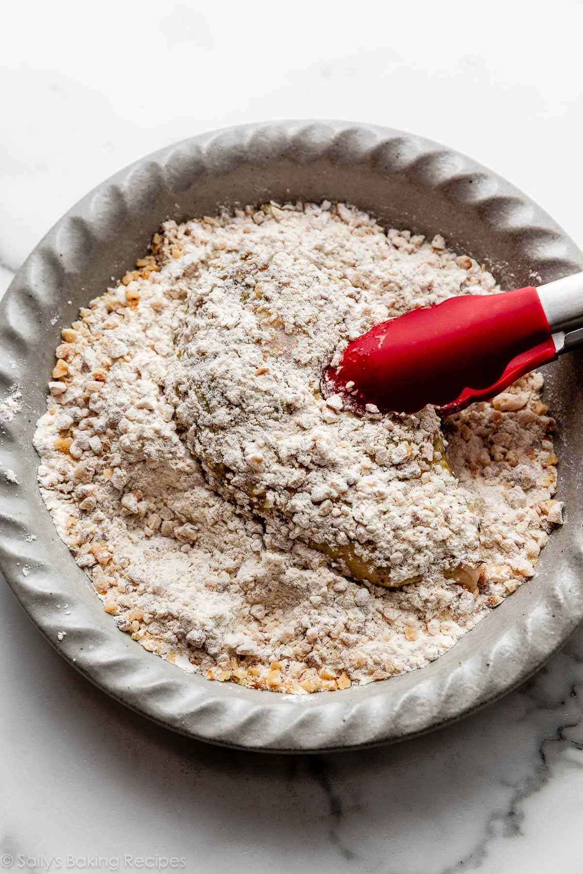 red tongs holding chicken breast in walnut and flour mixture.