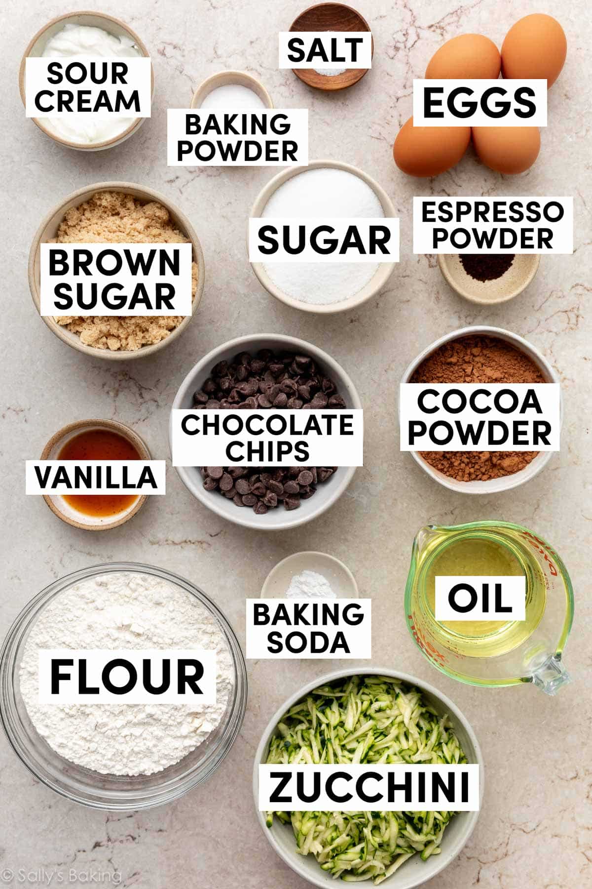 ingredients on marbled surface including chocolate chips, cocoa powder, oil, flour, eggs, espresso powder, brown sugar, and sour cream.