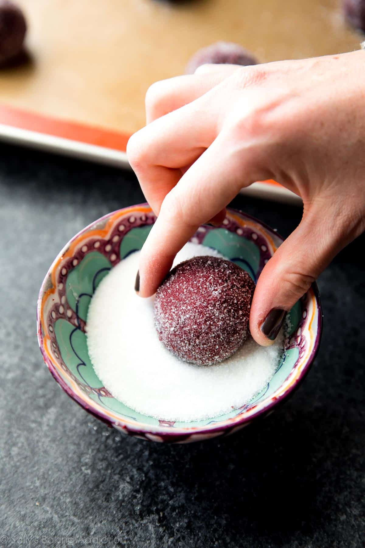hands rolling red velvet cookie dough ball into a bowl of sugar