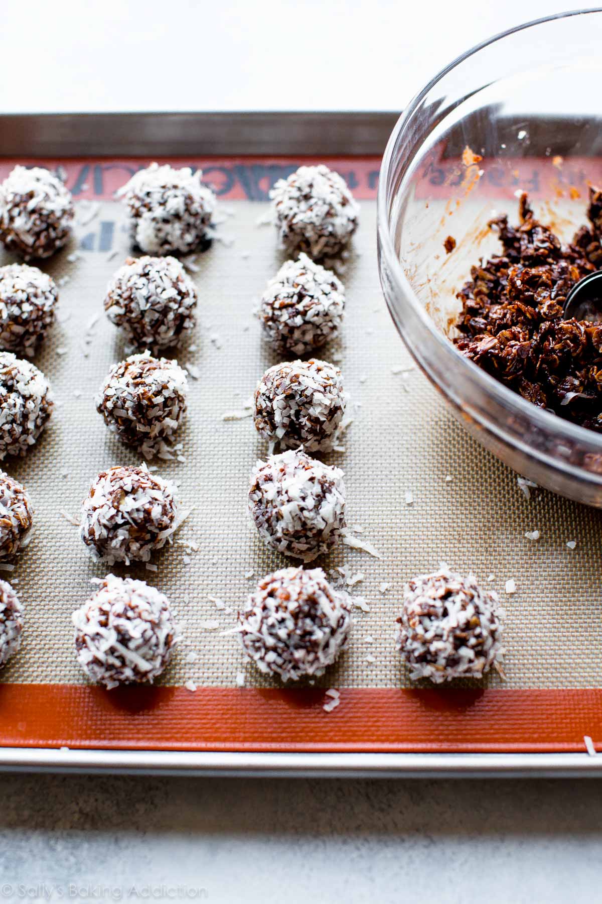 no-bake chocolate coconut snowballs on a baking sheet with a bowl of chocolate oat mixture