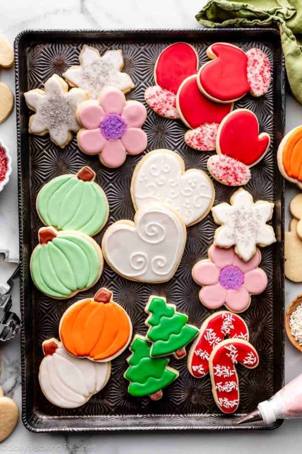 decorated sugar cookies on baking sheet including hearts, pumpkins, trees, mittens, and flowers.