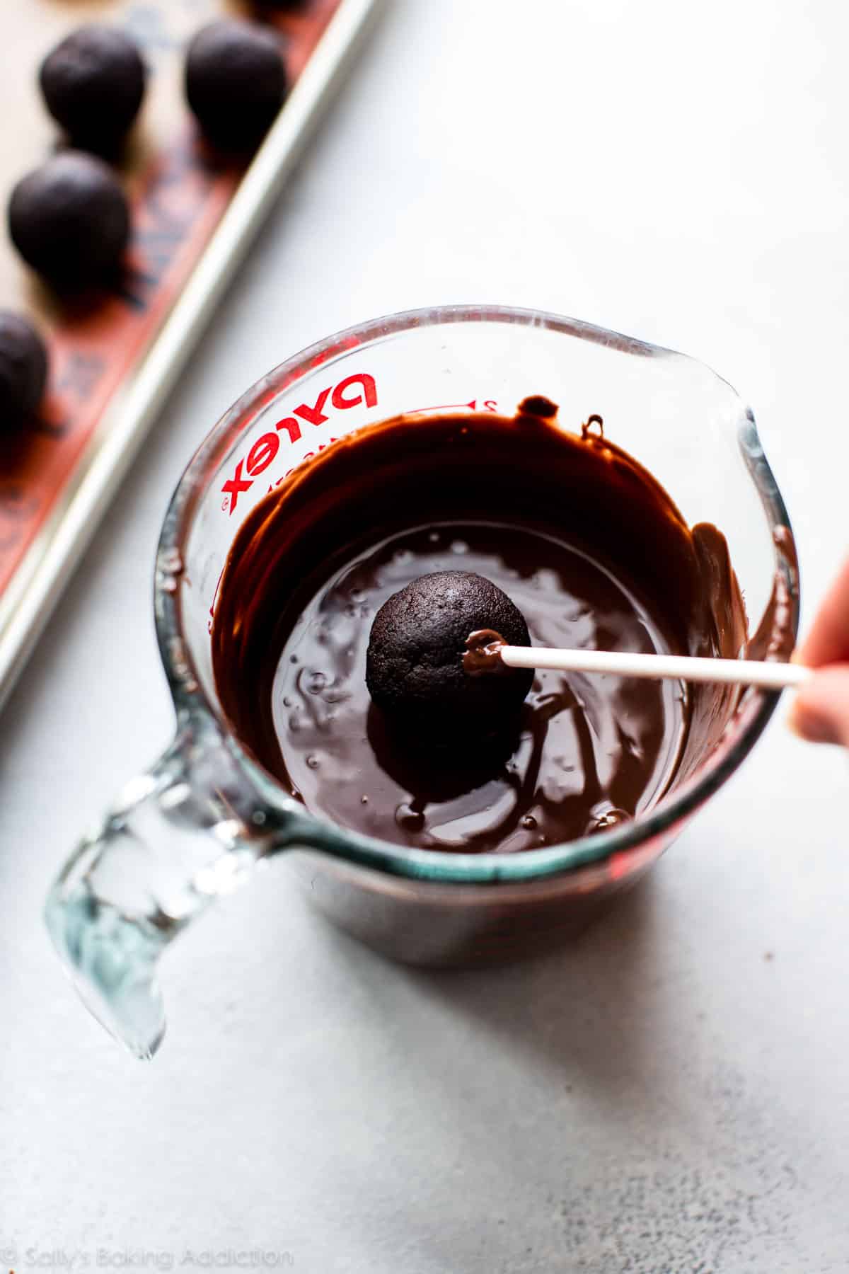 dipping a chocolate cake pop on a stick into chocolate coating in a glass measuring cup