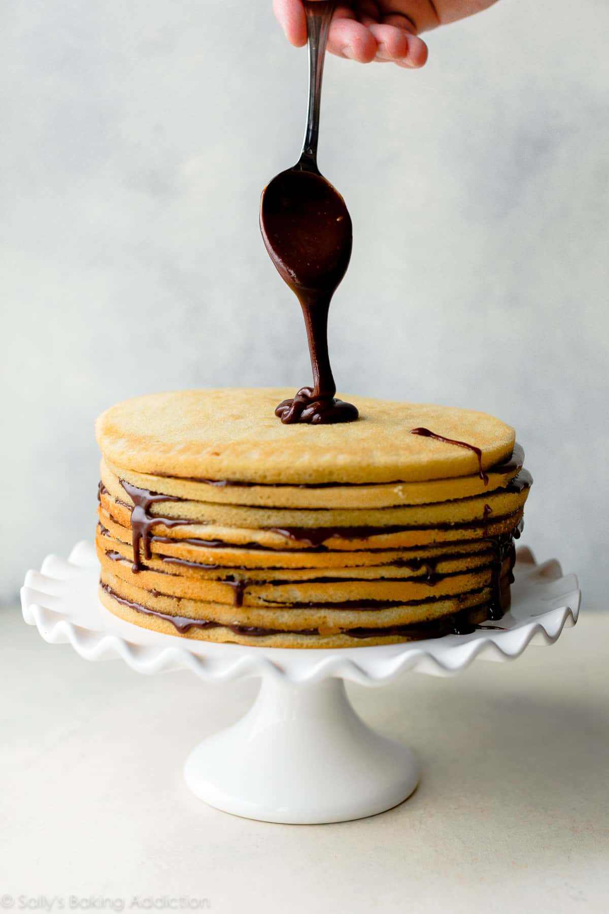 spooning chocolate fudge icing onto top of Smith Island Cake on a white cake stand