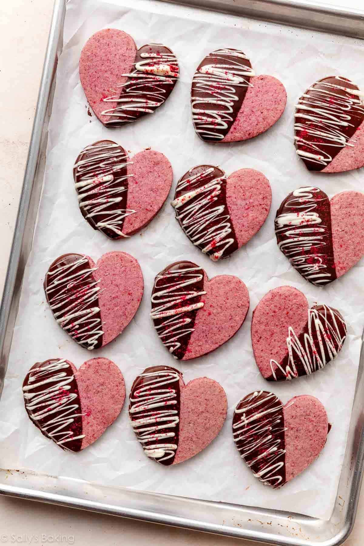 heart-shaped raspberry Valentine's Day cookies dipped in chocolate and drizzled with white chocolate on lined baking sheet.