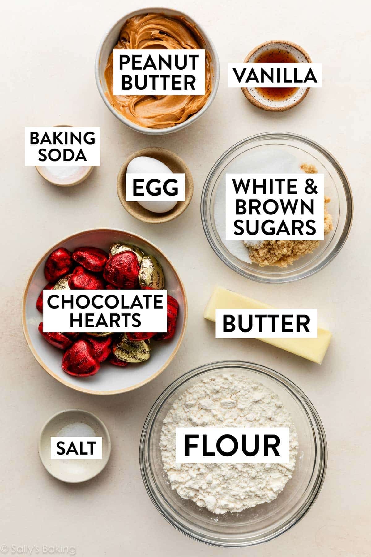 ingredients in bowls on beige surface including butter, sugar, egg, vanilla, flour, and wrapped chocolate heart candies.