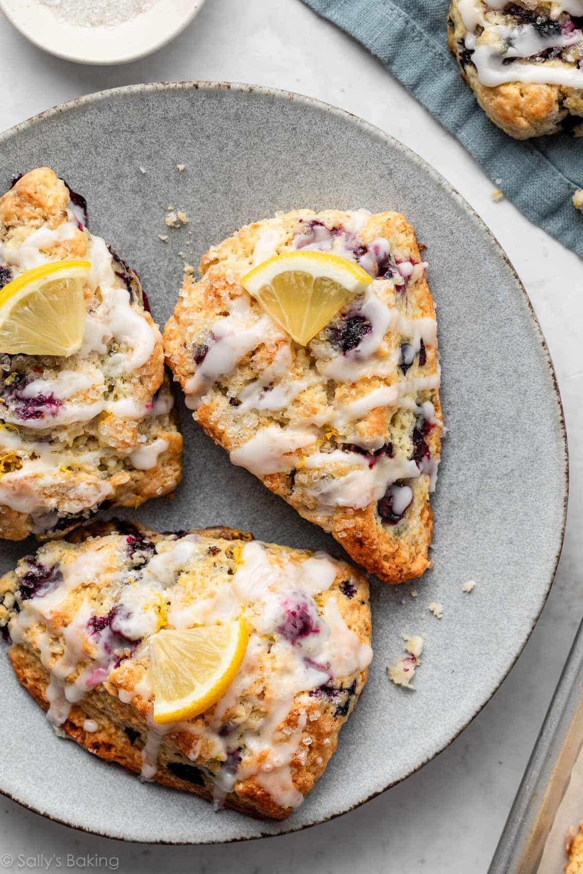 lemon blueberry scones with fresh lemon slices and icing on top on a blue-gray colored-plate.