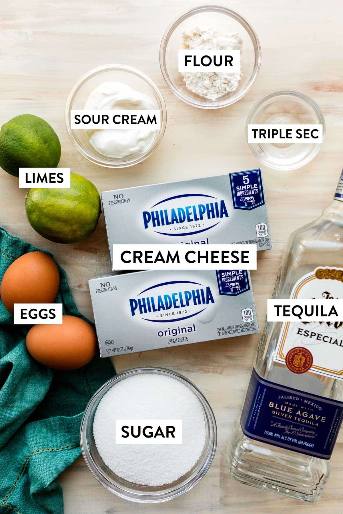 cream cheese, bottle of tequila, triple sec, flour, sour cream, limes, eggs, and sugar in bowl.