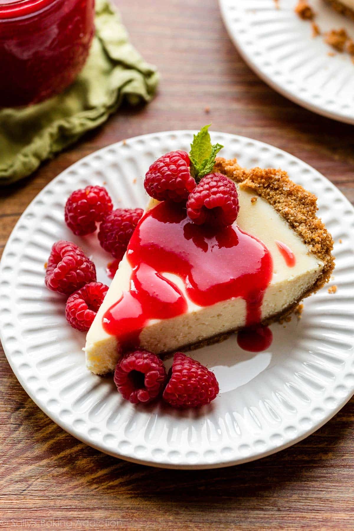 cheesecake slice on white plate with raspberry sauce and fresh raspberries on top.