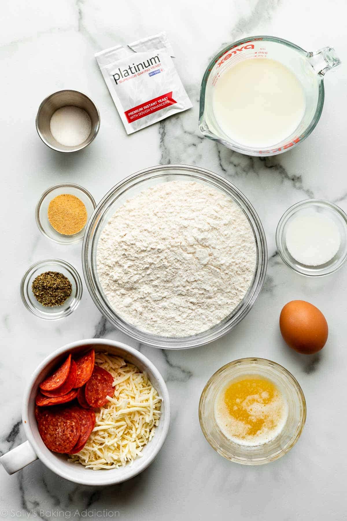 bread flour, milk, egg, sugar, melted butter, seasonings, pepperoni, cheese, and other ingredients on marble counter.