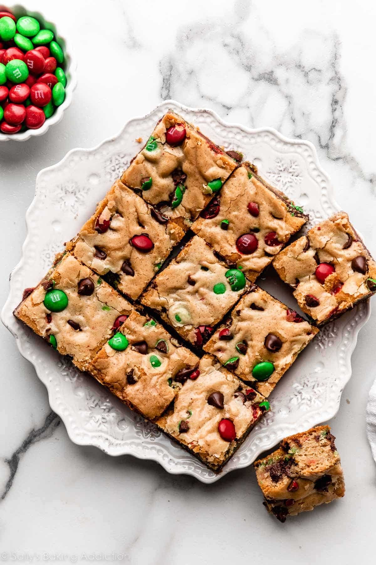 9 Christmas M&M cookie bars arranged on white snowflake-shaped plate.
