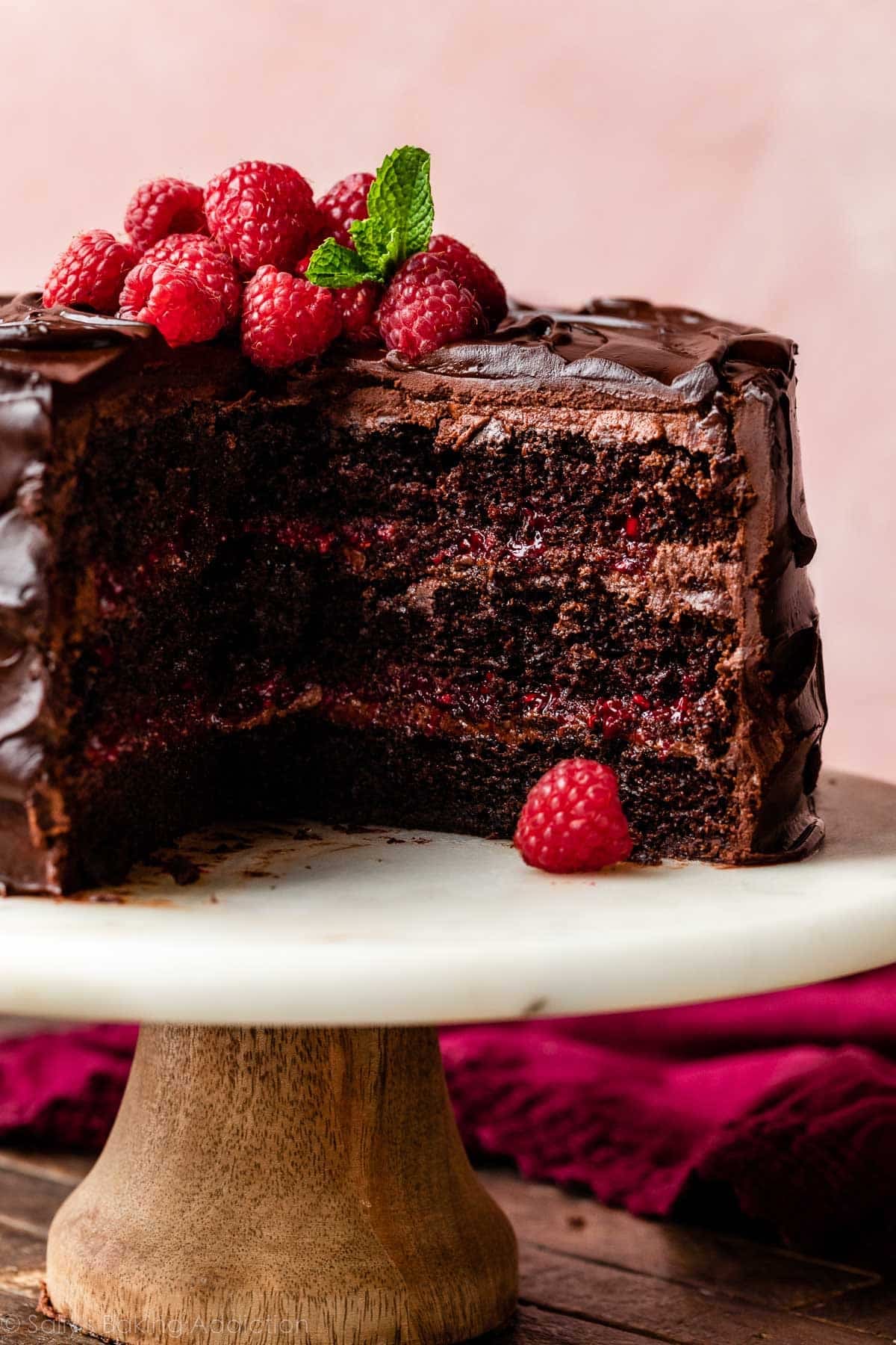 chocolate raspberry cake cut open with raspberries and mint on top sitting on a marble cake stand with pink backdrop behind it.