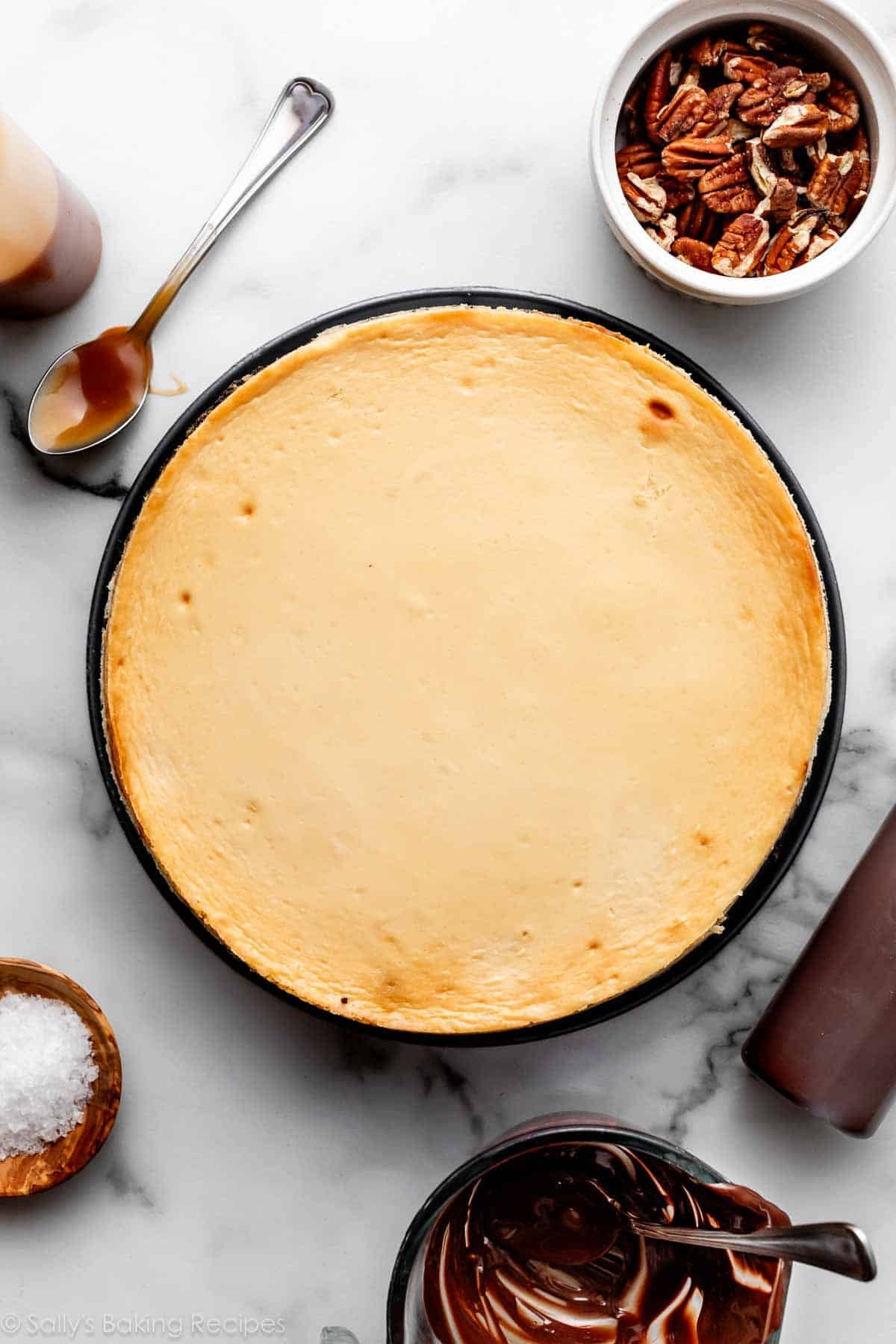 brown sugar cheesecake on marble counter with pecans, spoonful of caramel, sea salt, and melted chocolate around it.