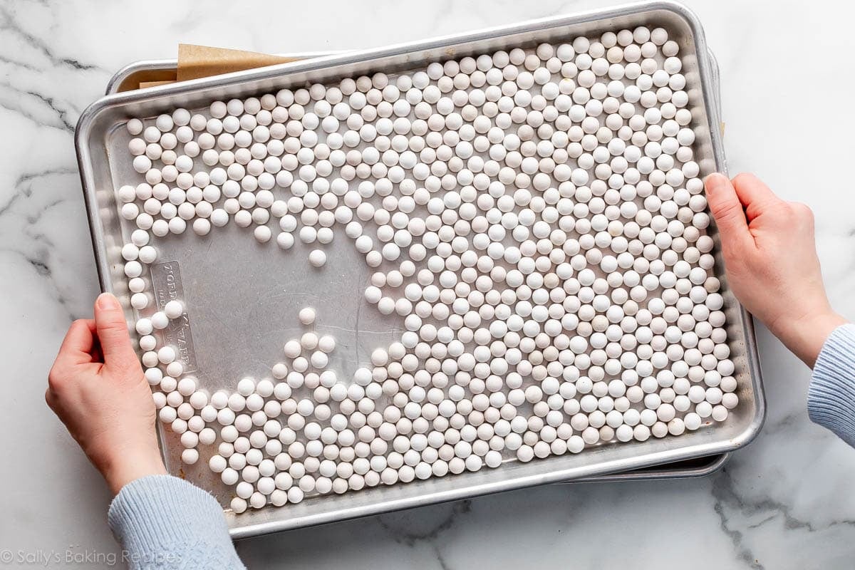 hands holding a baking sheet with white round pie weights on top.