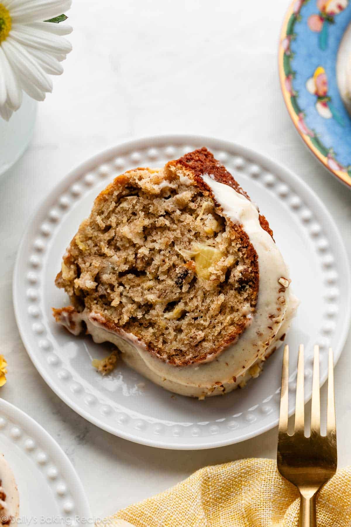 slice of hummingbird Bundt cake with cream cheese icing on white plate with gold fork.