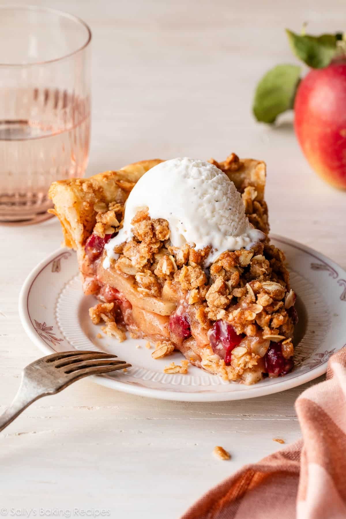 slice of apple cranberry crumb pie on white plate with vanilla ice cream on top and pink glass in the background.