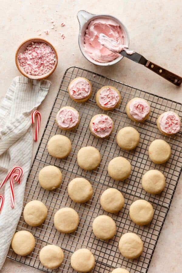 cookies on silver cooling rack with pink frosting and crushed candy canes in a bowl next to it.