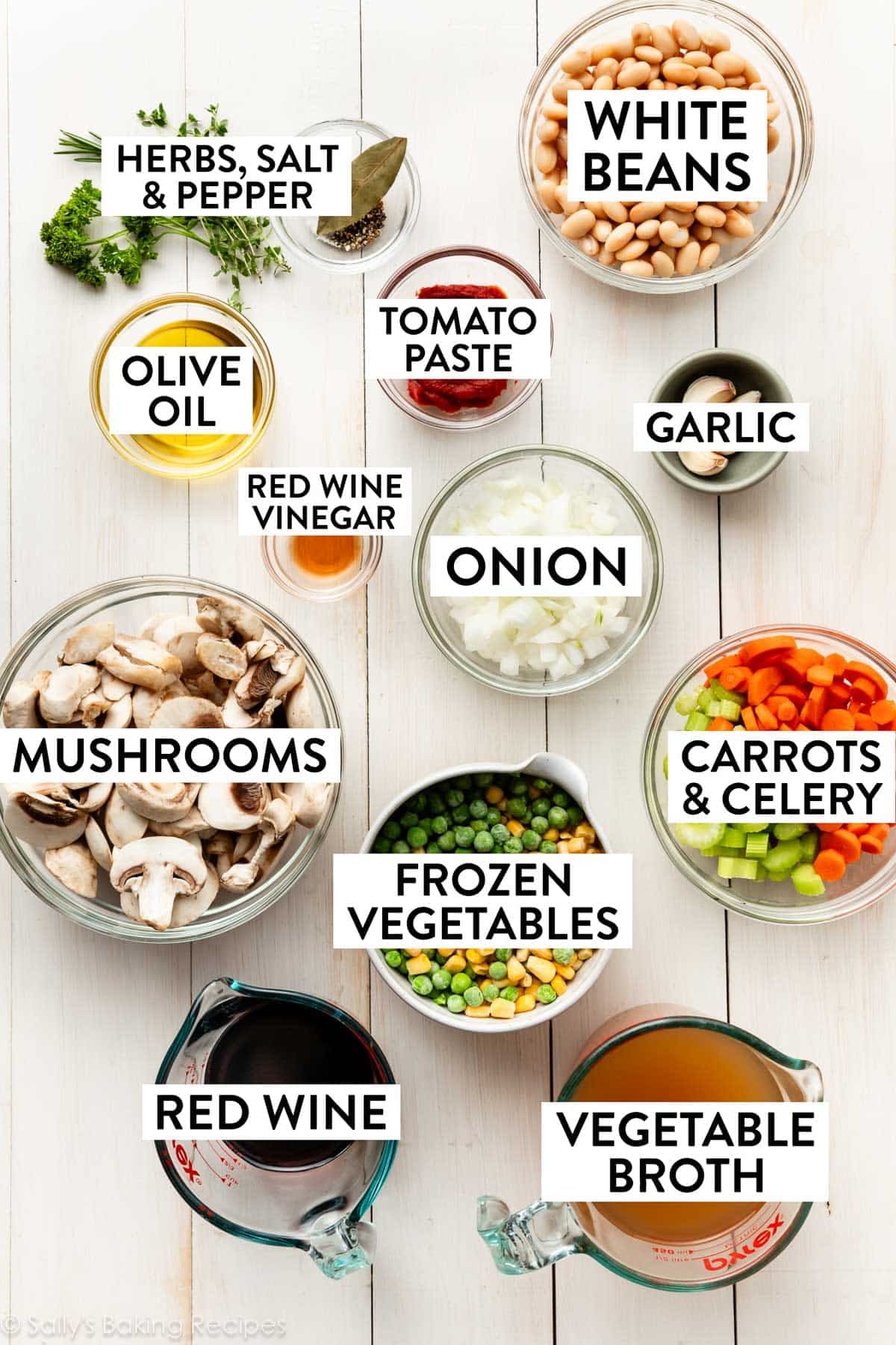 ingredients on white wooden surface including peas, carrots, celery, mushrooms, chopped onions, white beans, herbs, and red wine.