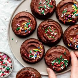 hand grabbing chocolate cookie with chocolate frosting and Christmas sprinkles off plate.