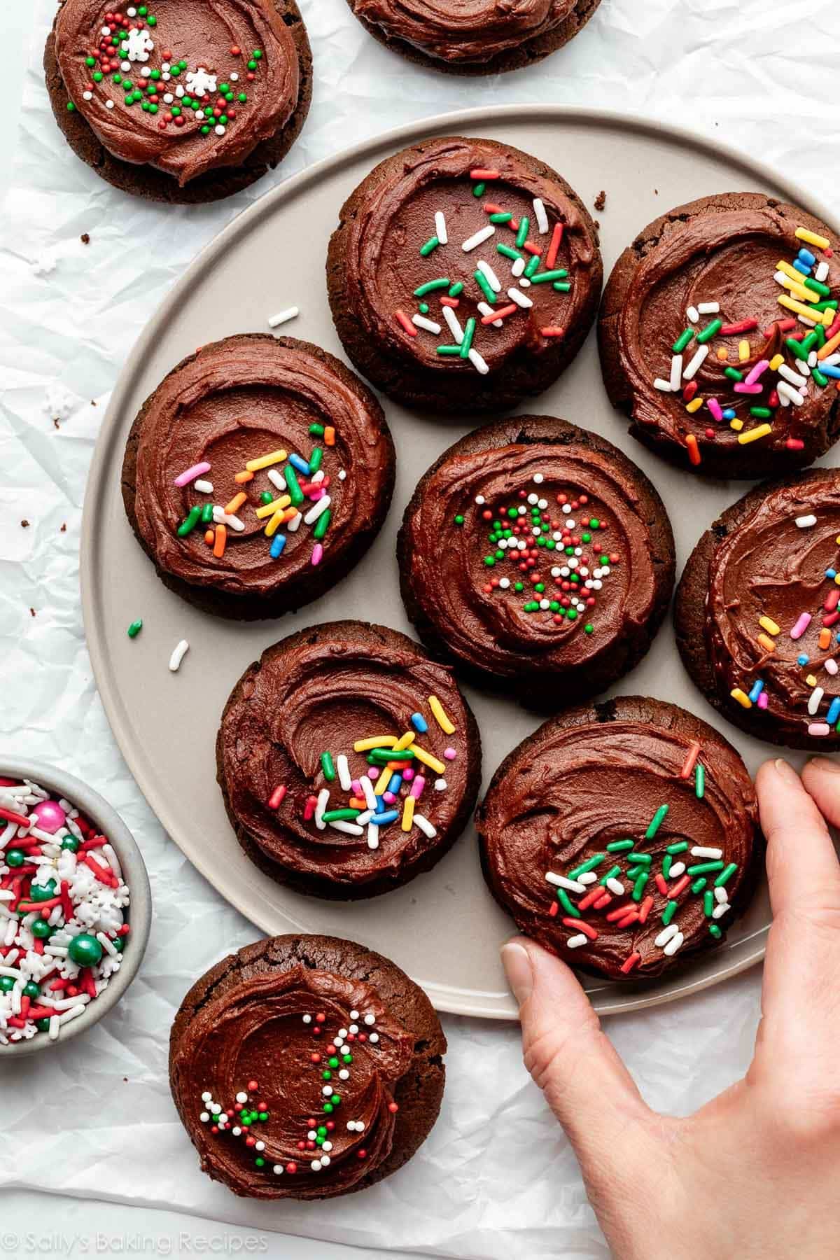 hand grabbing chocolate cookie with chocolate frosting and Christmas sprinkles off plate.