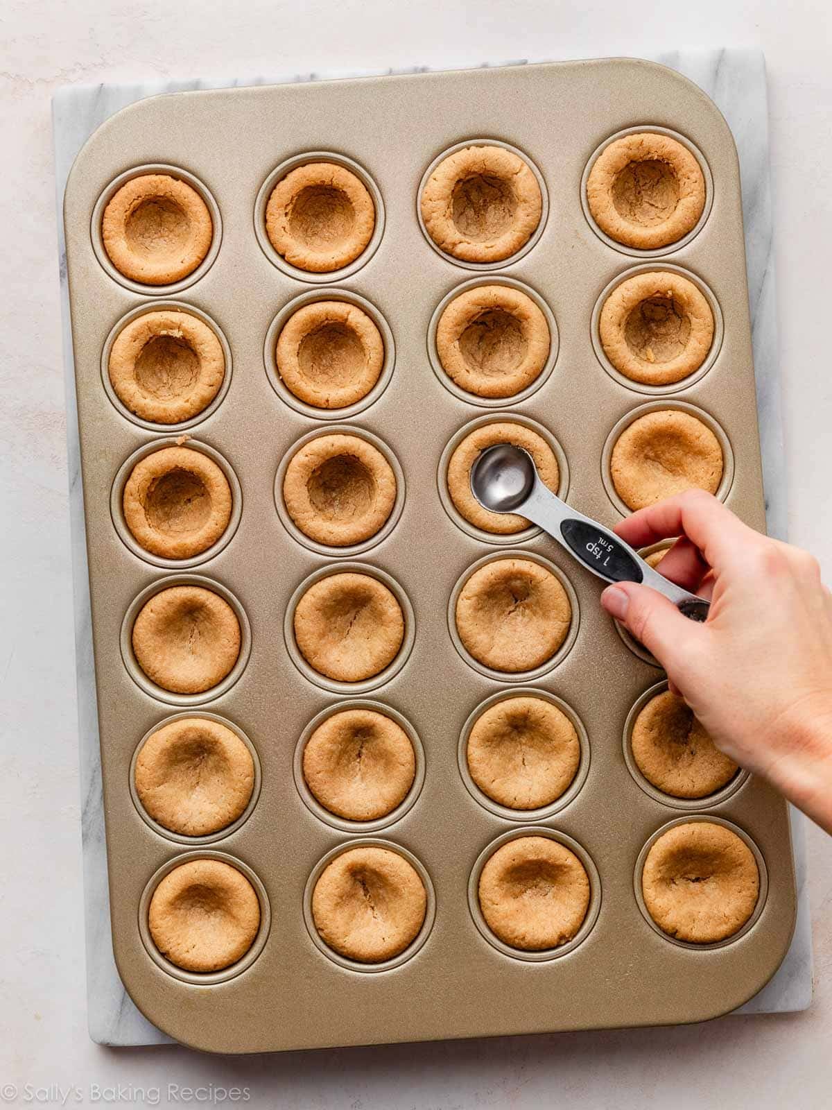 hand and measuring spoon making indents into peanut butter cookie cups in a mini muffin pan.