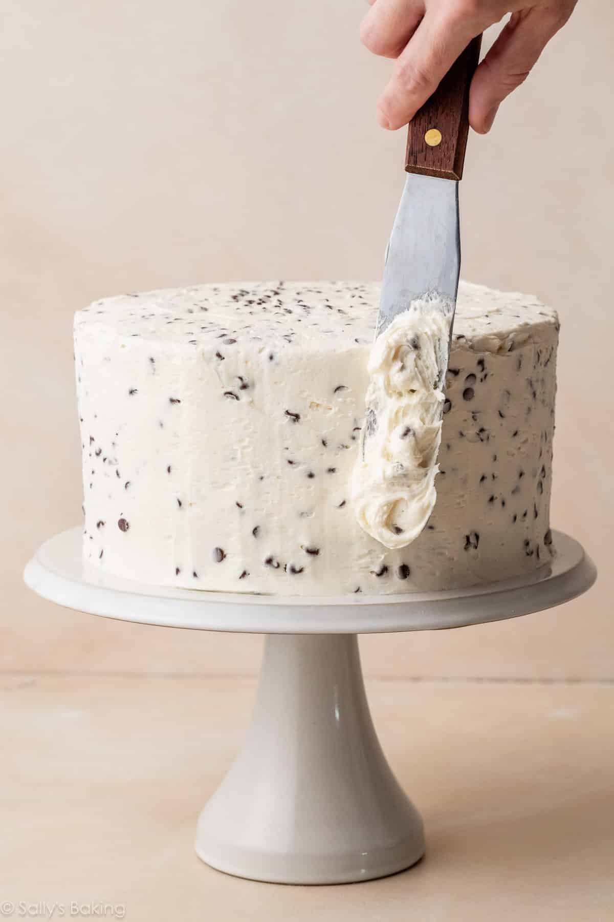 hand spreading frosting on layer cake on white cake stand.