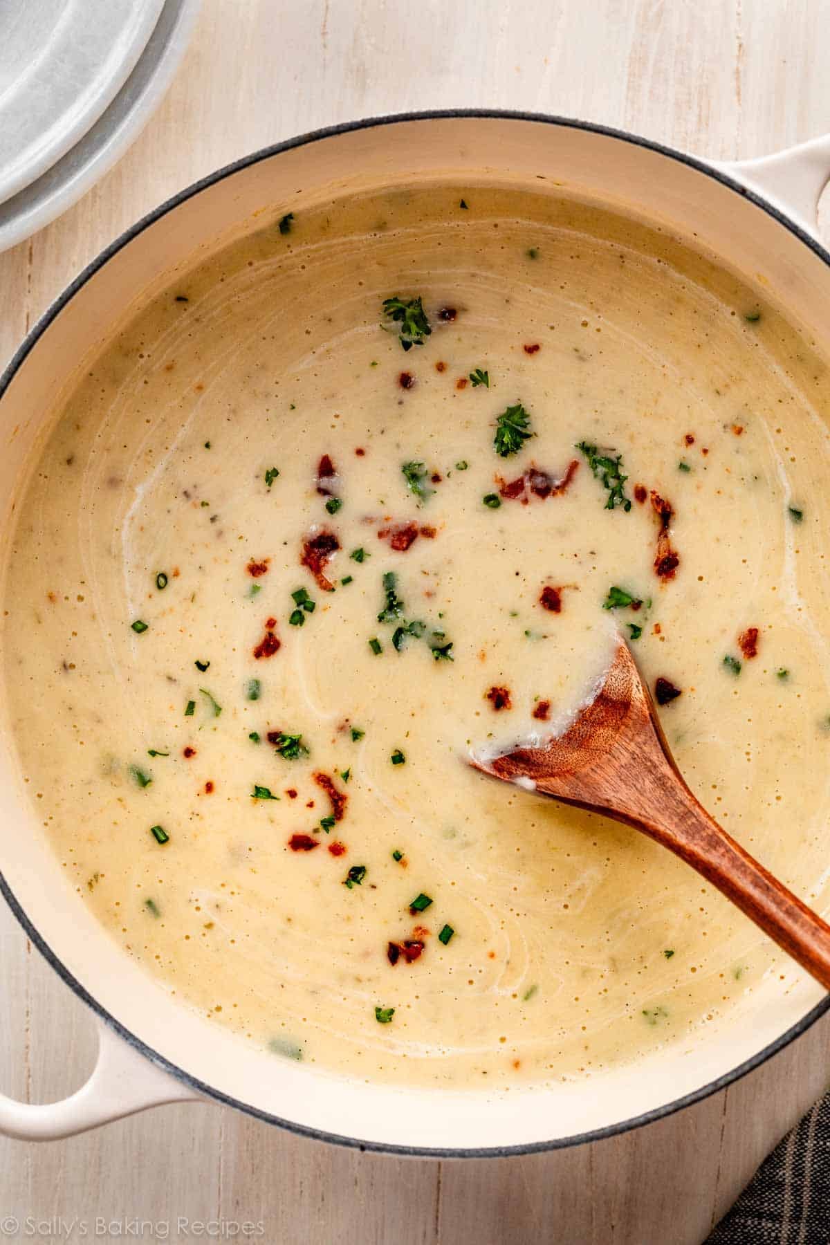 pot of cauliflower potato soup with wooden spoon topped with parsley, chives, and bacon.