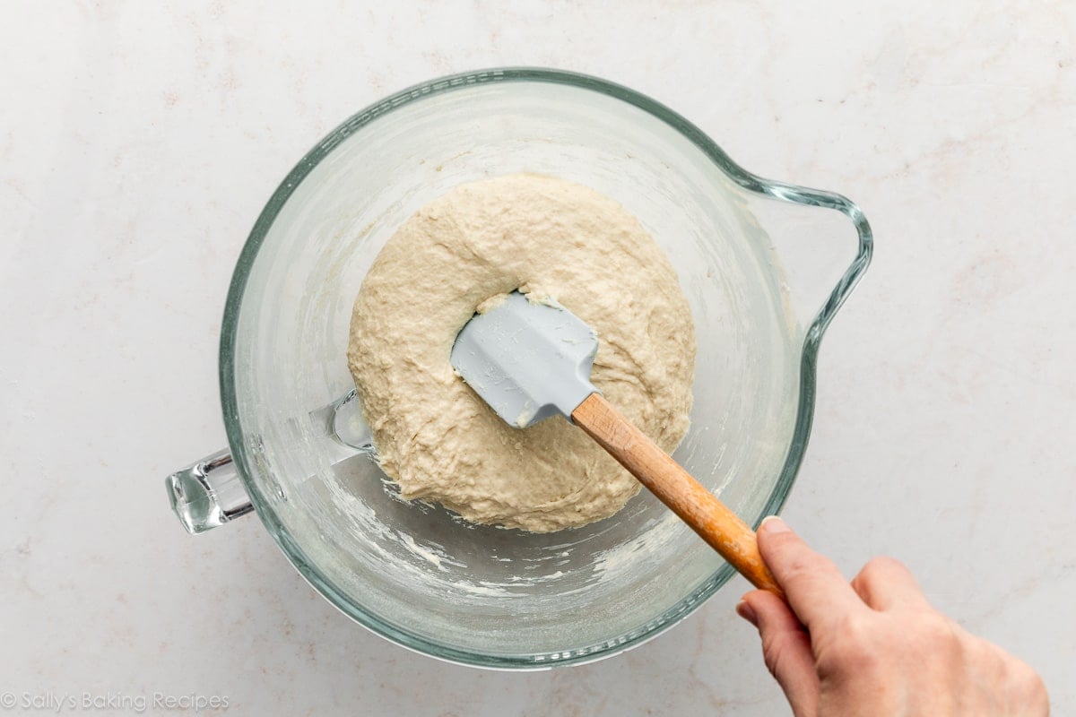 hand holding blue spatula on top of dough in glass bowl.