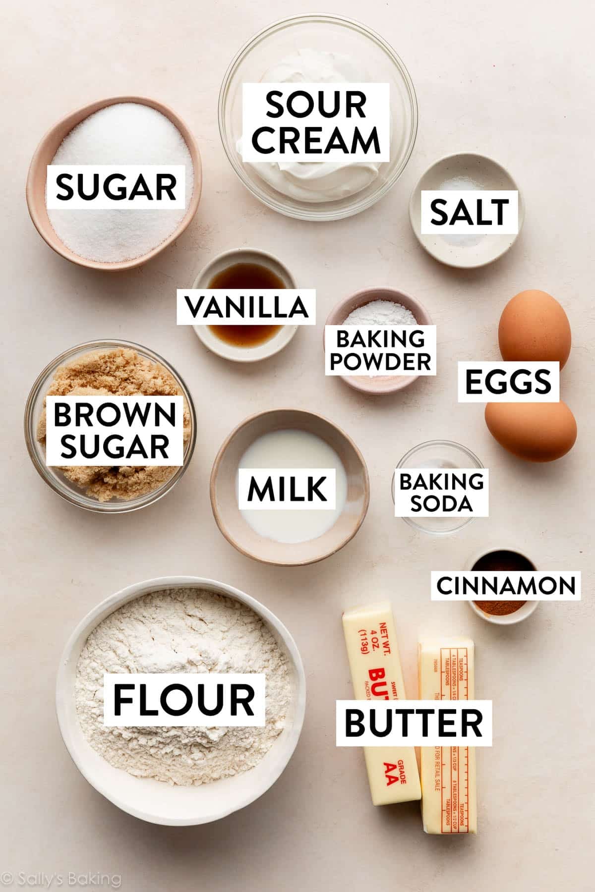 ingredients in bowls on beige surface including butter, sour cream, salt, sugar, vanilla, brown sugar, milk, and eggs.