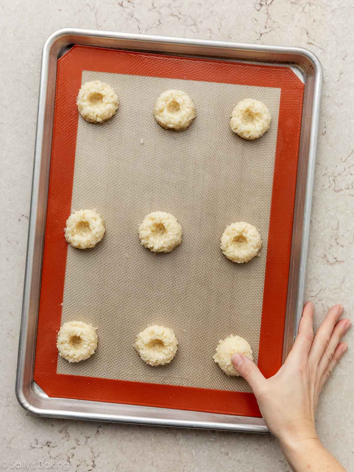 thumb pressing thumbprint in cookie dough on silicone baking mat-lined pan.